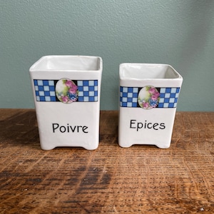 May include: Two white ceramic spice jars. Each jar has a blue and white checkered band with a floral design. One jar reads "Poivre" and the other reads "Epices". The jars are square and have a small foot.