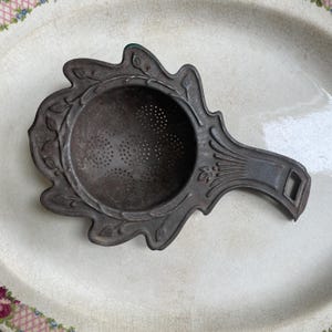May include: Vintage tea strainer with an ornate, leaf-like design. The metal strainer has a circular basket with small perforations for tea leaves. The handle has a diamond-shaped opening. The strainer is a dark, aged metal colour.