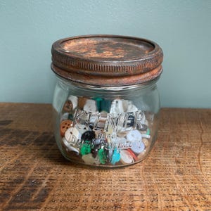 May include: A clear glass Kilner jar filled with assorted buttons. The jar has a rusty metal lid and the Kilner logo is visible on the glass. The buttons are various colours and sizes, including white, black, green, and red.