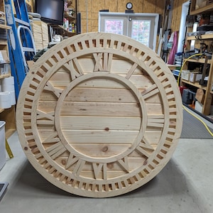 May include: Large, unfinished wooden clock face with Roman numerals. The clock is circular, made of light-colored wood, and features a detailed design with cut-out elements. The Roman numeral XII is prominently displayed at the top.