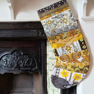 May include: A patchwork Christmas stocking with a variety of floral and geometric patterns in shades of yellow, gold, black, and white. The stocking is hanging from a white shelf above a fireplace.