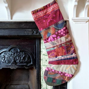 May include: A patchwork Christmas stocking in shades of burgundy, pink, and green, hanging from a gold hook. The stocking features various fabric patterns and textures, creating a festive and unique holiday decoration.