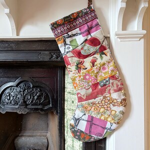 May include: A patchwork Christmas stocking with floral and geometric patterns in pink, red, orange, and green. The stocking hangs from a white mantelpiece above a black fireplace, a festive decoration.