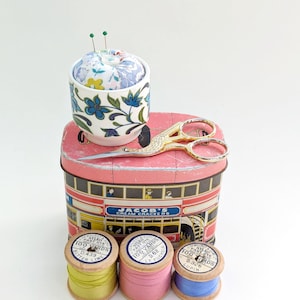 May include: A vintage sewing kit featuring a floral pin cushion, gold scissors, and spools of thread. The items are arranged on a pink tin with the text "JACOB'S CREAM CRACKERS". The thread spools are yellow, pink, and blue.