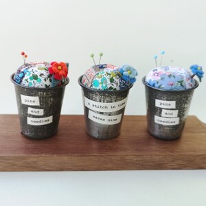 May include: Three small metal containers with fabric pincushions inside. Each container has a different saying on the side: "pins and needles", "a stitch in time saves nine", and "pins and needles".
