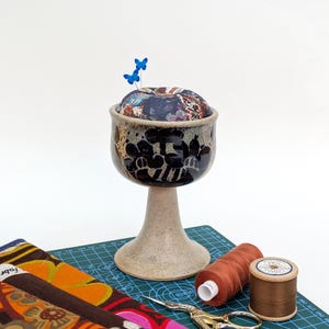 May include: A decorative ceramic pin cushion with a floral pattern, featuring two blue butterfly pins. The pin cushion is on a beige pedestal. Sewing items, including thread spools, scissors, and fabric, are arranged on a green cutting mat.