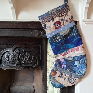 May include: A patchwork Christmas stocking in shades of blue, beige, and red, featuring floral and patterned fabrics. The stocking is suspended near a fireplace, with a loop for hanging.
