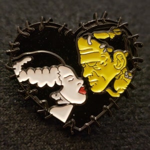 May include: Black and white enamel pin in the shape of a heart with a stitched border. The heart is divided in half, with a yellow Frankenstein's monster on the right and a white Bride of Frankenstein on the left. Both figures are looking at each other.