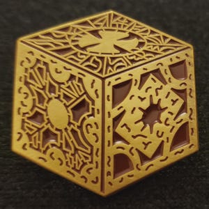 May include: A gold-colored, cube-shaped pin with intricate geometric patterns and a cross design on the top. The pin has a detailed, embossed appearance with brown accents, creating a three-dimensional effect. The pin is likely a collectible or decorative item.