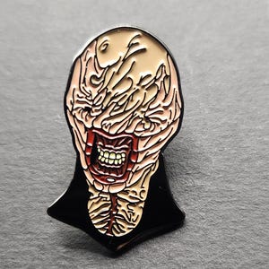 May include: Enamel pin featuring a grotesque, pale face with a wide, open mouth revealing teeth. The design includes a black collar and a detailed, textured appearance, with red and beige tones.