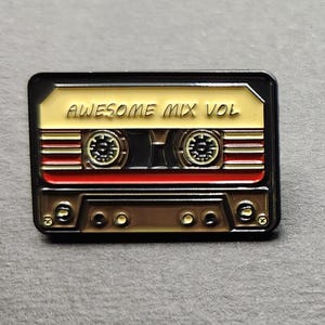 May include: A rectangular pin featuring a retro cassette tape design. The pin has a gold and brown color scheme with red accents. The text "AWESOME MIX VOL" is displayed on the top of the pin.