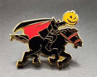 May include: A gold-toned metal enamel pin featuring a headless figure in a black suit with a red-lined cape, riding a black horse. The figure holds a yellow jack-o'-lantern with a carved face. The horse has a red bridle and black body.