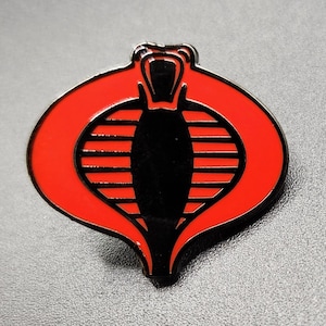 May include: A red and black enamel pin featuring the Cobra symbol. The pin has a red outer shape with a black cobra head and vertical lines in the center. The pin is a collectible item.