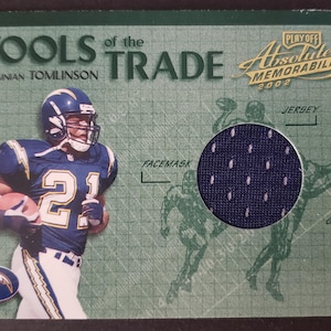 May include: A trading card featuring LaDainian Tomlinson in a blue and white Chargers uniform. The card includes the text "TOOLS of the TRADE" and "PLAYOFF Absolute Memorabilia 2002". A circular inset shows a fabric swatch.