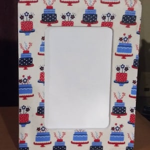 May include: A white picture frame with a red, white, and blue patterned border featuring a cake design. The frame has a white mat and a clear glass front.