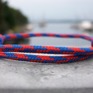 Puede incluir: Un brazalete de cuerda trenzada rojo y azul con un nudo en un extremo.