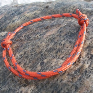 May include: A close-up of a red and blue rope bracelet tied in a knot. The bracelet is lying on a grey and brown textured surface.
