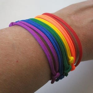 Könnte beinhalten: Eine Reihe von Armbändern aus Regenbogenfarbenem Seil. Die Armbänder sind in einem Farbspektrum angeordnet: rot, orange, gelb, grün, blau und lila. Die Armbänder sind mit Knoten versehen und werden am Handgelenk getragen.