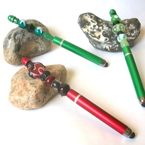 Könnte beinhalten: Drei Eingabestifte mit dekorativen Glasperlen an den oberen Enden. Zwei Stifte haben grüne Metallgehäuse und ein Stift hat ein rotes Metallgehäuse. Jeder Stift ist mit einer einzigartigen Anordnung bunter Glasperlen und einer schwarzen Spitze versehen.