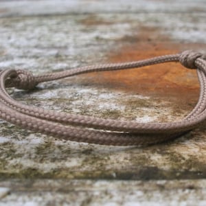Pode incluir: Uma pulseira de corda trançada de cor castanha com um fecho de nó. A pulseira é feita de um material tecido durável e foi concebida para ser usada no pulso. A pulseira é mostrada numa superfície desgastada e texturizada.
