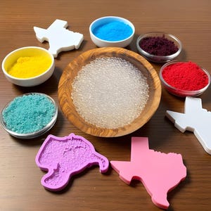 May include: A set of six small bowls filled with colorful powders, including red, blue, yellow, and purple. A wooden bowl filled with clear beads sits in the center. Six silicone molds in the shape of the state of Texas are arranged around the bowls. The molds are in various colors, including pink, purple, and white.