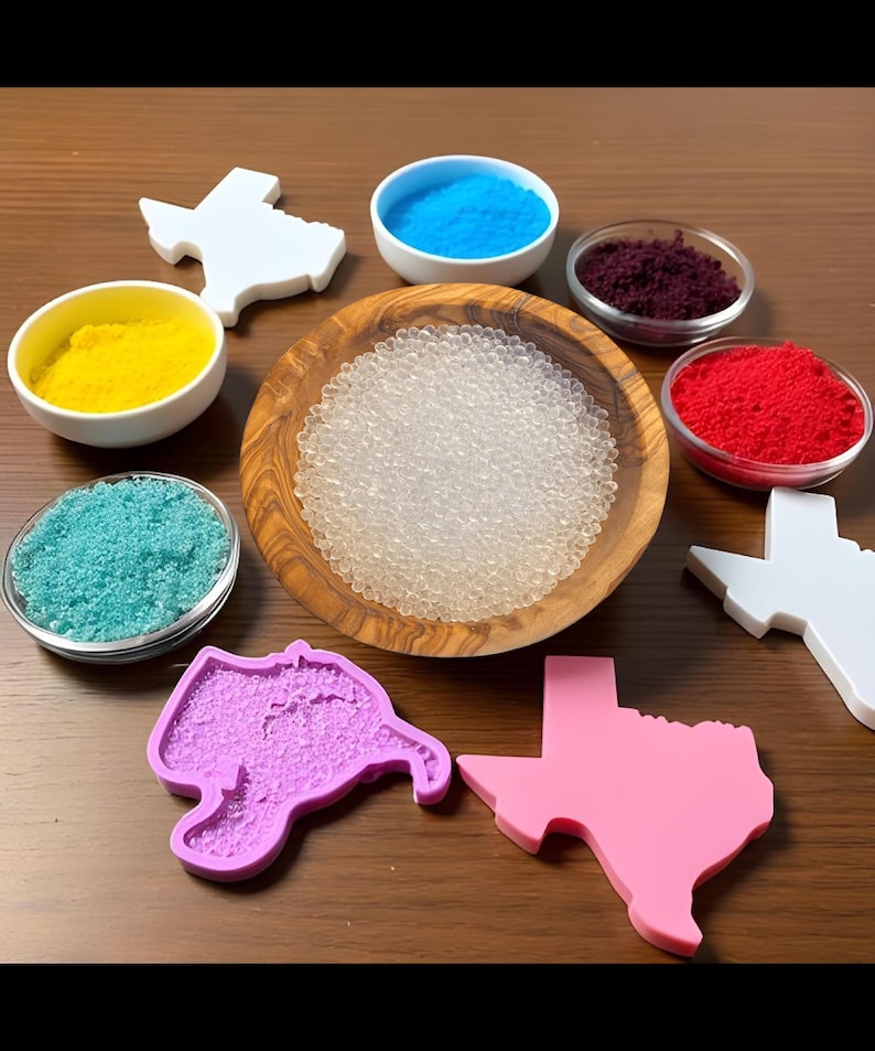 May include: A set of six small bowls filled with colorful powders, including red, blue, yellow, and purple. A wooden bowl filled with clear beads sits in the center. Six silicone molds in the shape of the state of Texas are arranged around the bowls. The molds are in various colors, including pink, purple, and white.