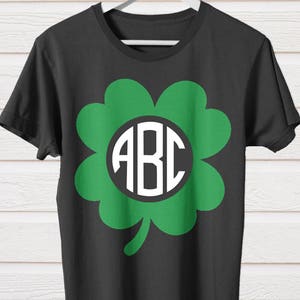 Puede incluir: Camiseta negra con un diseño de trébol de cuatro hojas verde y un círculo blanco con las letras "ABC" en su interior.