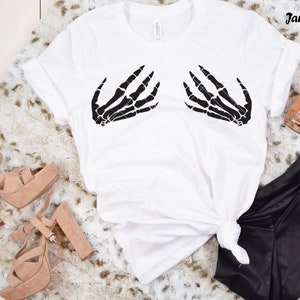 May include: White t-shirt with a black skeleton hand graphic on the front. The shirt is laying on a white faux fur surface with a pair of brown heeled sandals and black pants.