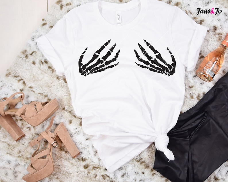 May include: White t-shirt with a black skeleton hand graphic on the front. The shirt is laying on a white faux fur surface with a pair of brown heeled sandals and black pants.