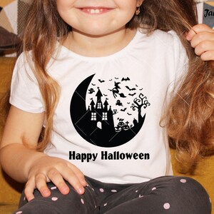 May include: A white t-shirt with a black silhouette of a crescent moon, a haunted house, a witch flying on a broomstick, bats, a ghost, a tree, and pumpkins. The text "Happy Halloween" is printed below the moon.