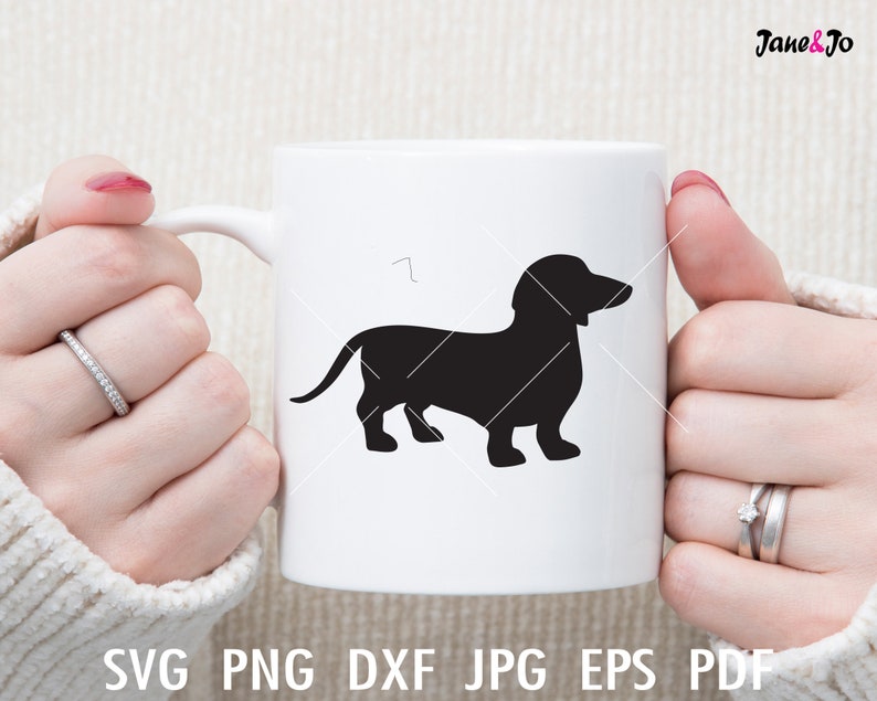 Może przedstawiać: Biały ceramiczny kubek z czarną sylwetką psa rasy dachshund. Kubek jest trzymany przez dłonie osoby.