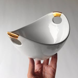 May include: A white ceramic bowl with an unusual, asymmetrical shape. The bowl has gold-coloured accents on the handles, which have circular openings. The bowl is held up, showing its smooth, glossy surface. This decorative bowl is ideal for serving.