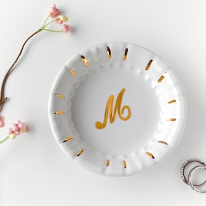 May include: White ceramic ring dish with gold accents and a gold letter "M" in the center. The dish has a scalloped edge with cut-out details. A sprig of pink flowers and two rings are next to the dish.
