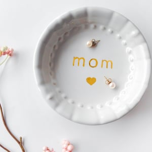 May include: A white ceramic trinket dish with a scalloped edge, featuring the gold word "mom" and a gold heart. Two pearl earrings rest on the dish. Pink and white decorative sprigs are in the background.