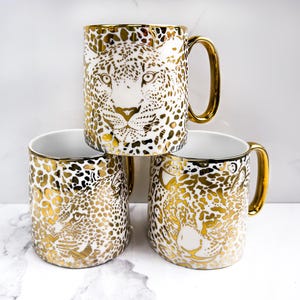 Könnte beinhalten: Drei weiße Keramikbecher mit goldenem Leopardenmuster. Jeder Becher hat einen goldenen Henkel und ein goldenes Leoparden-Gesichtsmuster. Die Becher sind übereinander gestapelt.