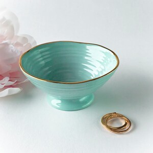 May include: A light blue ceramic bowl with a gold rim. The bowl has a pedestal base and a textured interior. Two gold rings are next to the bowl. A pink flower is in the background.