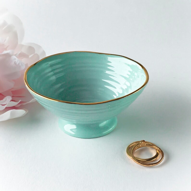 May include: A light blue ceramic bowl with a gold rim. The bowl has a pedestal base and a textured interior. Two gold rings are next to the bowl. A pink flower is in the background.