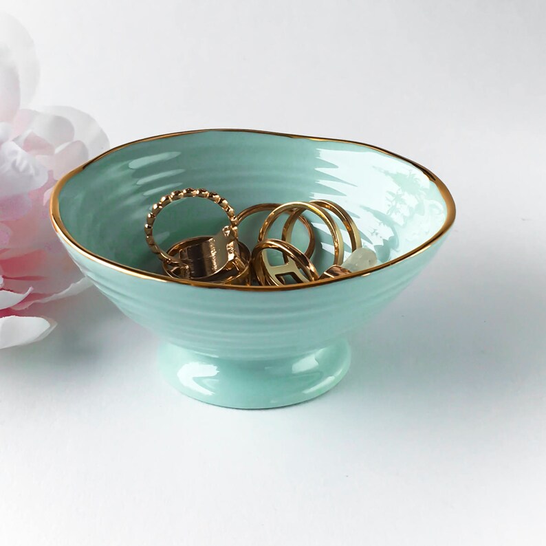 May include: A light blue ceramic jewelry dish with a gold rim, holding several gold rings. The dish has a pedestal base and a ribbed texture. A pink flower is partially visible in the background.