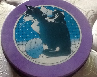 May include: A purple round tin with a black and white illustration of two cats. The larger cat is sitting with its back to the viewer and the smaller cat is sitting on its back. The illustration is on a blue background with white polka dots.