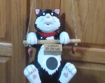 May include: A black and white cat figurine hanging from a string with a wooden sign that reads "Lunch is on me".