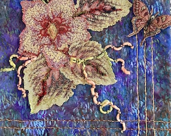 May include: A close-up of a fabric art piece with a floral design. The piece features a pink and yellow flower with green leaves and a butterfly in the upper right corner. The background is a blue and purple fabric with a textured pattern.