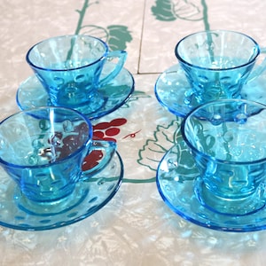 May include: Four matching vintage blue glass teacups and saucers. Each cup has a handle and sits on a matching saucer. The set is made of transparent blue glass with a textured design.