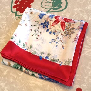 May include: A square scarf featuring a white background with a floral pattern in shades of red, blue, and green. The scarf has a solid red border along two edges. The fabric appears smooth and lightweight, folded to display the intricate botanical print.