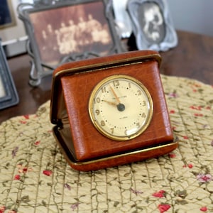 May include: A vintage brown leather travel alarm clock with a gold-tone bezel and a cream-colored face. The clock is open, revealing the time. The clock is sitting on a floral patterned surface.