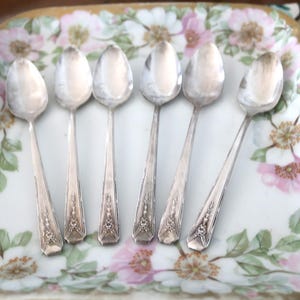 May include: Six silver spoons arranged on a floral-patterned serving dish. The spoons have a decorative design on the handles. The dish features a white background with pink and white flower illustrations and a gold trim.