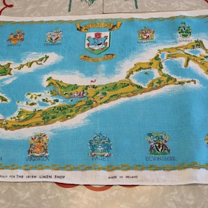May include: A vintage linen tea towel featuring a map of Bermuda in shades of blue, yellow, and green. The towel includes the names of several parishes and is bordered with a decorative gold trim. The text "DESIGNED BY DECORAMA EXPRESSLY FOR THE IRISH LINEN SHOP" is printed on the bottom.