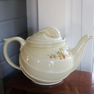 May include: Cream-colored ceramic teapot with a lid and a curved handle and spout. The teapot has a spiral design and a small floral pattern on the side. The teapot is sitting on a wooden surface.