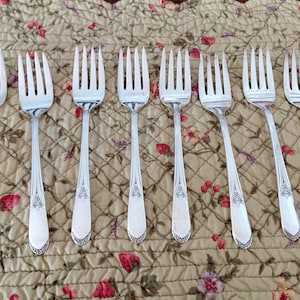 May include: A collection of eight silver forks with ornate handles, arranged on a floral patterned tablecloth. The forks have four tines and a decorative design on the handle.