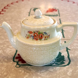 May include: A cream-colored ceramic teapot featuring a raised basket-weave texture across the body and lid. The center of the teapot is decorated with a colorful floral transfer print of orange poppies and small blue flowers, complete with a handle and spout.