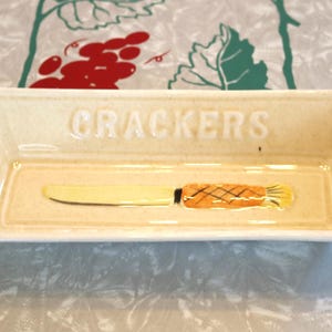 May include: A white ceramic cracker dish with a knife and crackers design. The dish has the word "CRACKERS" written on the front.